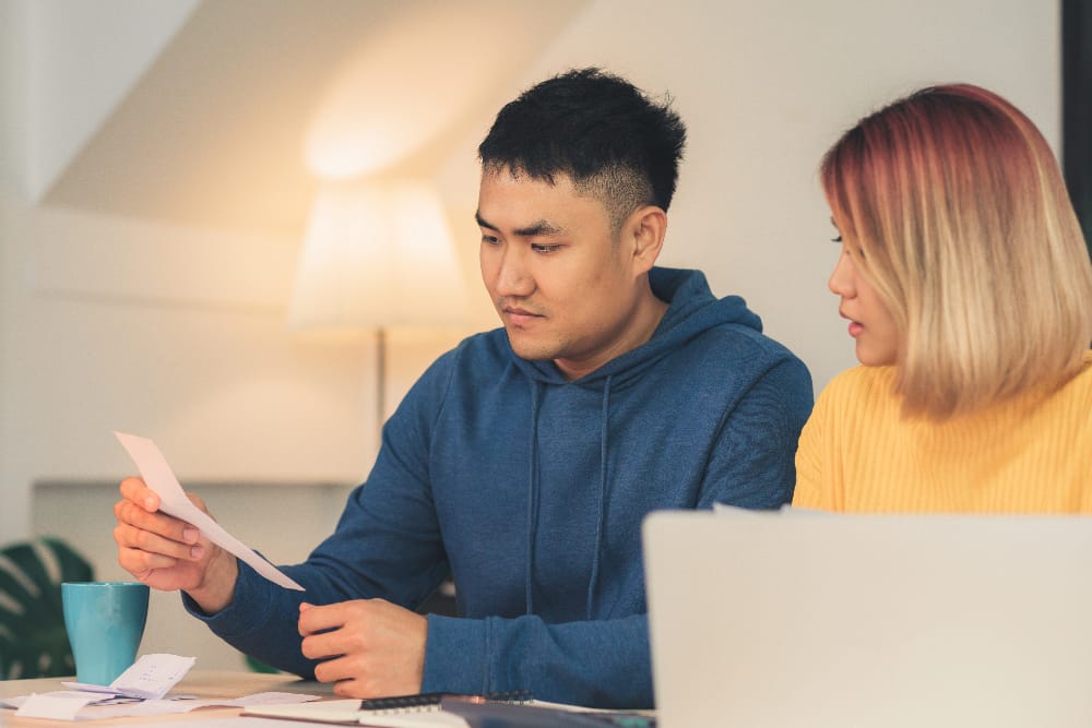A man in a blue hoodie and a woman in a yellow sweater sit at a table reviewing medical billing documents. Papers, a notebook, and a laptop are spread out as they discuss the No Surprises Act under the warm glow of a lamp in the background. - KindestMind