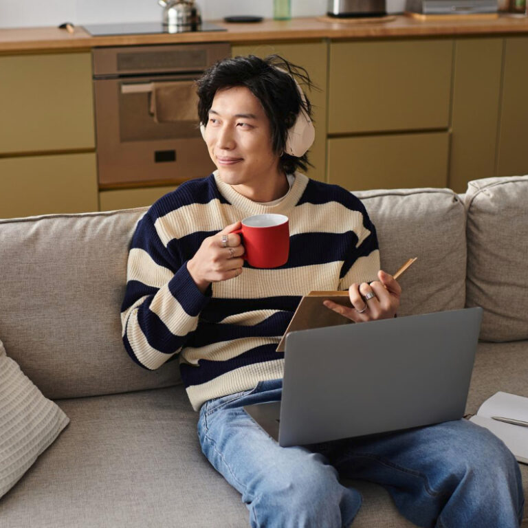 A person sits on a couch with a laptop on their lap, holding a red mug and a notebook. Wearing headphones and a striped sweater, they smile and look to the side—practicing self-accommodations for preventing burnout. The background shows a modern kitchen. - KindestMind