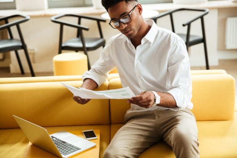 A man wearing glasses and a white shirt sits on a yellow couch, reviewing medical bills in his hand. An open laptop and a smartphone are on the couch beside him. - KindestMind