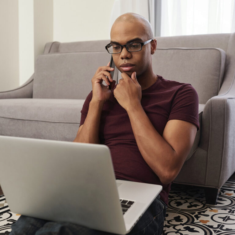 A man wearing glasses and a maroon t-shirt sits on the floor in front of a couch, looking at a laptop screen while talking on his phone, appearing thoughtful as he rests his hand on his chin, possibly reviewing assessments for Autism Spectrum Disorder. - KindestMind