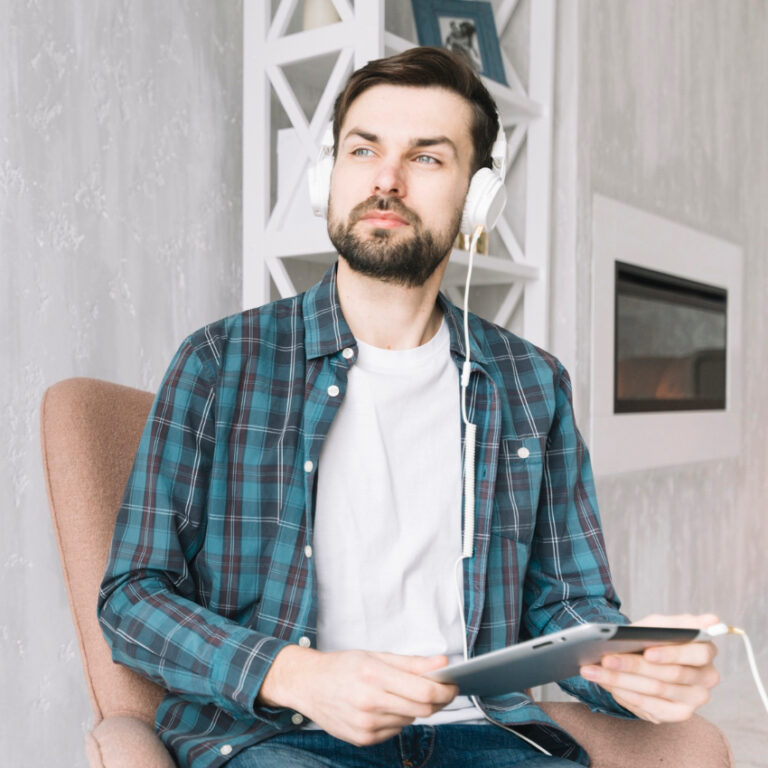 A bearded man in a plaid shirt sits in a chair, wearing headphones and holding a tablet used for assessments. He looks thoughtfully to the side. The background features a white shelf and a fireplace. - KindestMind