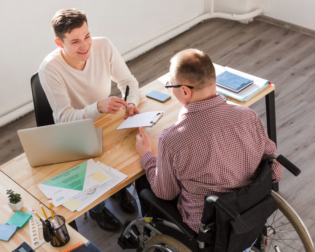 Two men sit across a desk; one is in a wheelchair. They are smiling and talking, with paperwork, a laptop, and documents on the desk in a bright office space, as they discuss details related to a Disability Assessment. - KindestMind