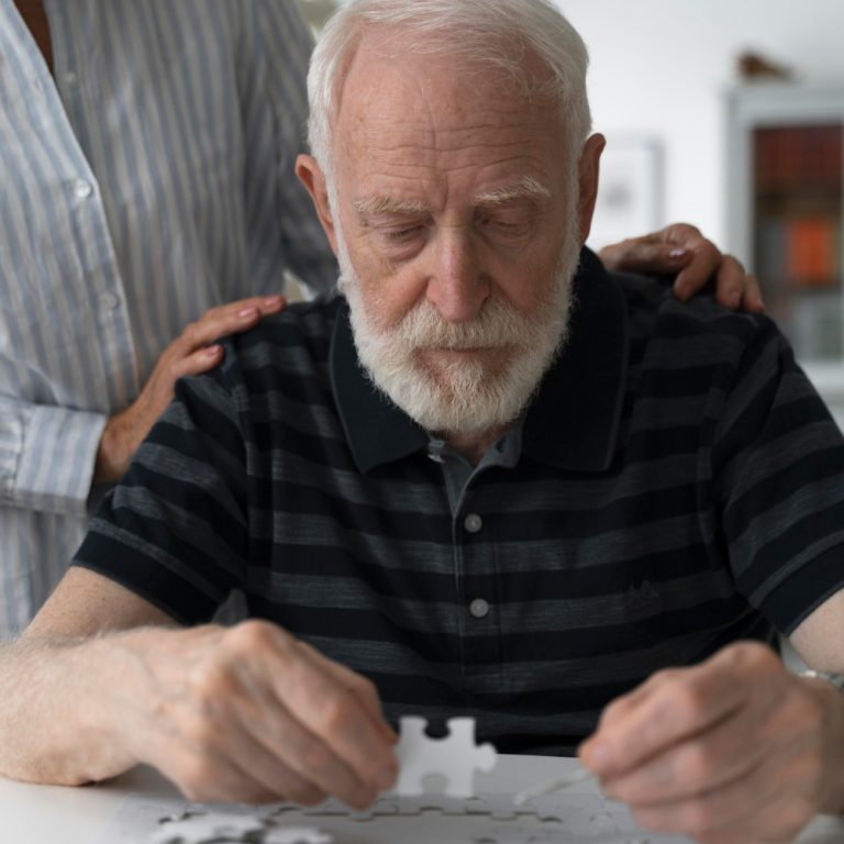 An elderly man with white hair and a beard focuses on assembling a puzzle. He sits at a table while another person rests comforting hands on his shoulders, offering support—an intimate moment often seen during dementia care. - KindestMind