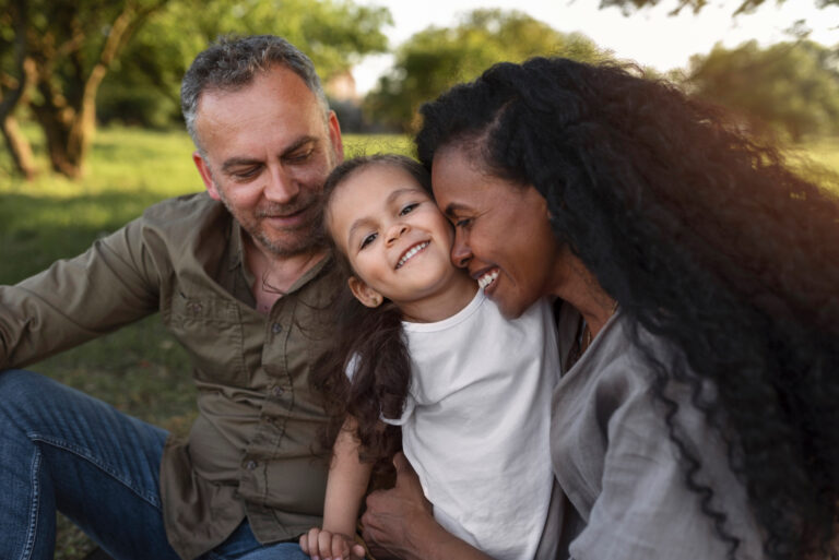 A man and woman sit on the grass with a child, reflecting the joy of supporting families through adoption. - KindestMind