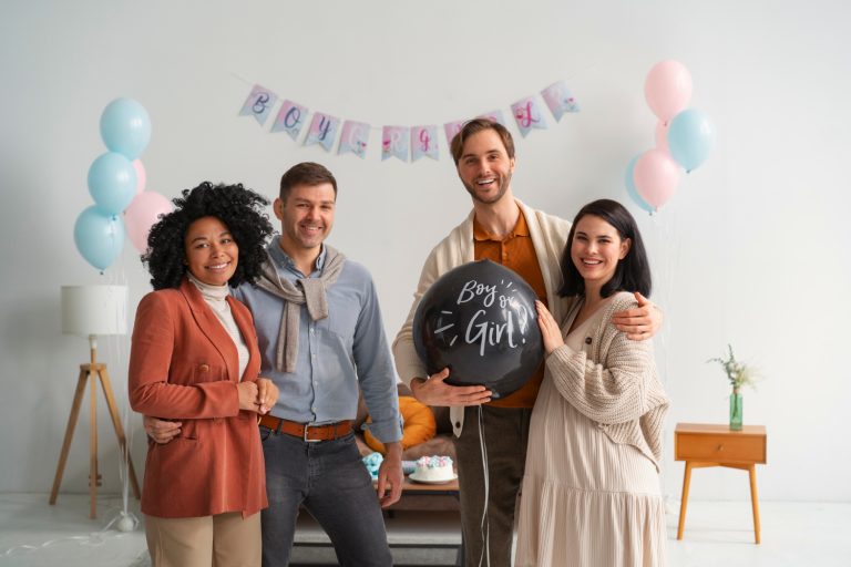 Four smiling adults stand together at a gender reveal party, holding a black balloon that reads Boy or Girl? Pink and blue decorations fill the background, celebrating their journey with independent surrogacy. - KindestMind