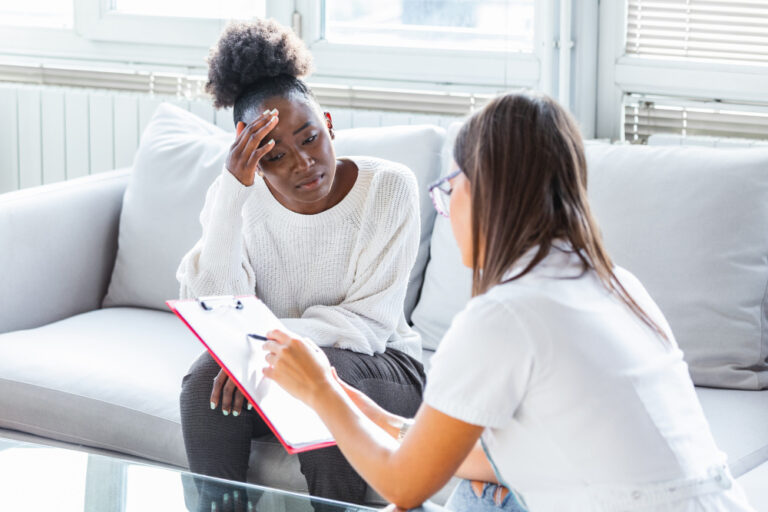 A woman sits on a couch, looking concerned with her hand on her forehead, while another woman with a clipboard talks to her—possibly during an Immigration Psychological Evaluation for VAWA applicants. - KindestMind