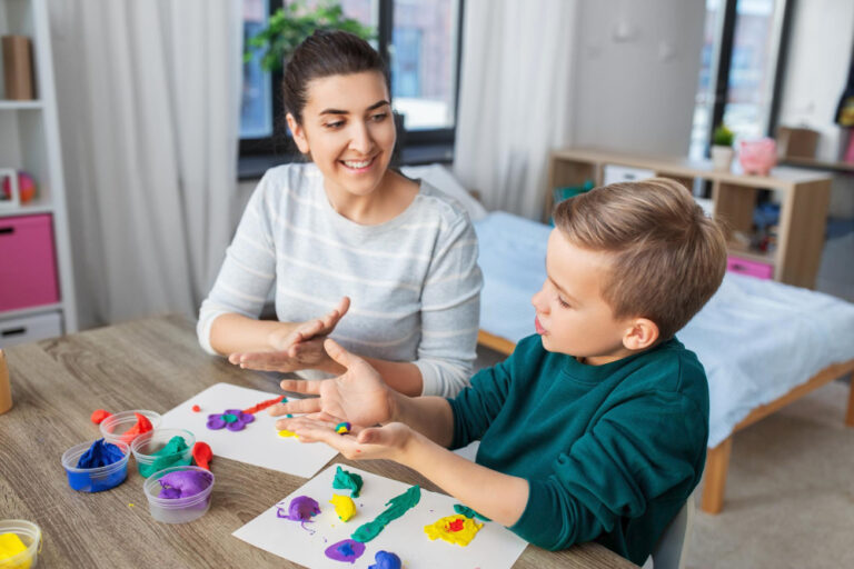 A woman and a young boy sit at a table indoors, smiling and painting with colorful finger paints on paper—showcasing autism strengths as they bond over creativity. Art supplies and finished artwork are spread out in front of them. - KindestMind