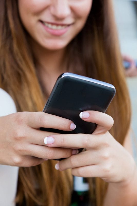 A young woman with long brown hair is smiling while holding and using a smartphone. Her fingernails are manicured with a French tip design. Only the lower half of her face and hands are visible. - KindestMind