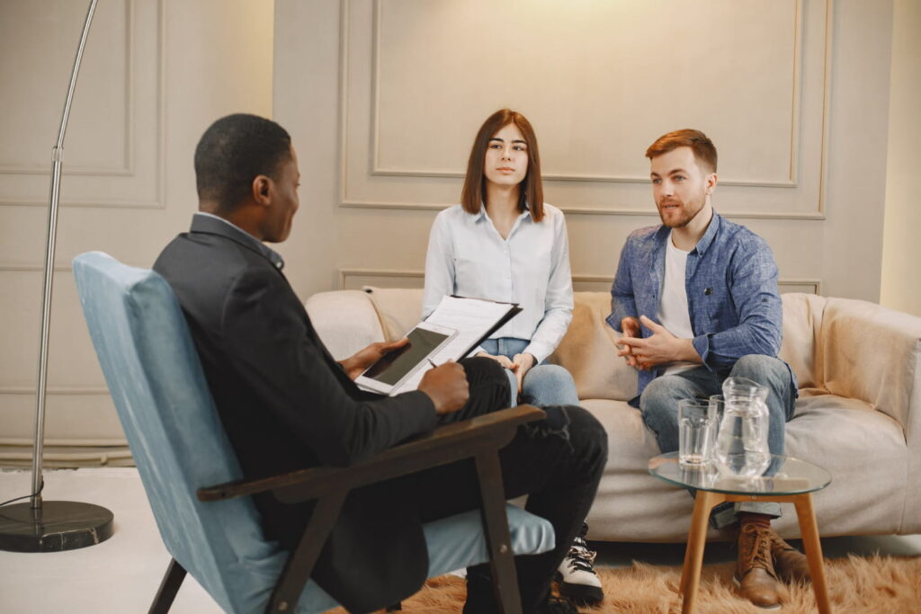 A man and woman sit on a couch talking to a therapist, who holds a clipboard. They appear to be having a counseling or therapy session in a modern, comfortable room, possibly as part of Independent Psychological Evaluations (IME) designed to assess their mental health needs. - KindestMind