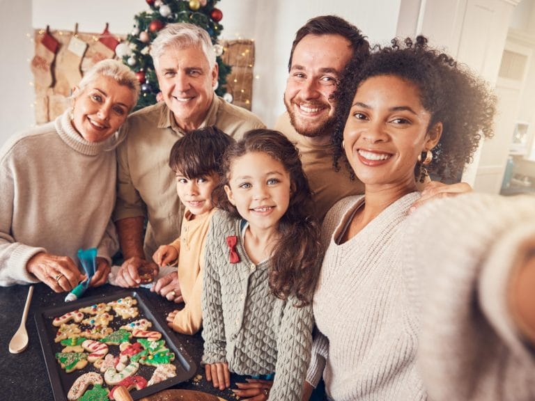 A multigenerational family smiles for a selfie in a festive kitchen, gathered around a tray of decorated holiday cookies, celebrating the holidays together with a Christmas tree and stockings in the background. - KindestMind