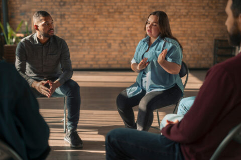 Home 5 A woman is speaking and gesturing with her hands in a support group setting focused on substance use assessments, while three others, including a man sitting nearby, listen attentively in a sunlit room with brick walls. - KindestMind