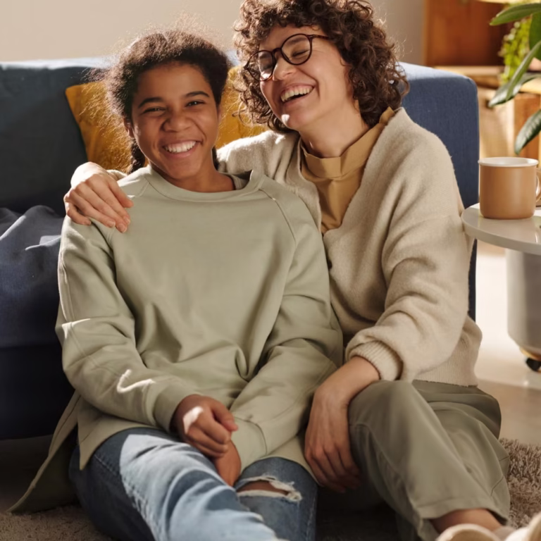 Two people sit on the floor in front of a blue sofa, smiling and hugging. One wears a light green top, the other beige. A table with a mug and book is beside them, as sunlight streams into the cozy room—perfect for quiet Juvenile Justice Evaluations. - KindestMind