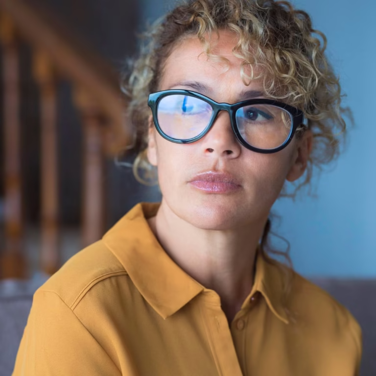 A woman with curly blonde hair and large black glasses wearing a mustard yellow shirt sits indoors, looking thoughtfully to the side—perhaps contemplating risk assessments. A wooden staircase is blurred in the background. - KindestMind