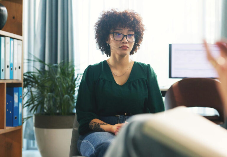 A woman with curly hair and glasses sits thoughtfully in an office, engaged in a conversation that suggests a Psychological Assessment or understanding Psychological Testing. Shelves and a computer are visible in the background. - KindestMind