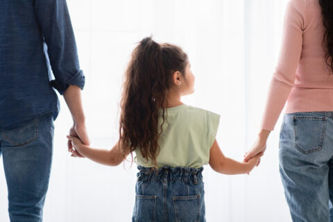 Home 4 A child with long curly hair stands between two adults, holding their hands—an image that reflects the closeness at the heart of any child custody or parental fitness evaluation, all seen from behind in casual clothes against a bright, softly lit background. - KindestMind