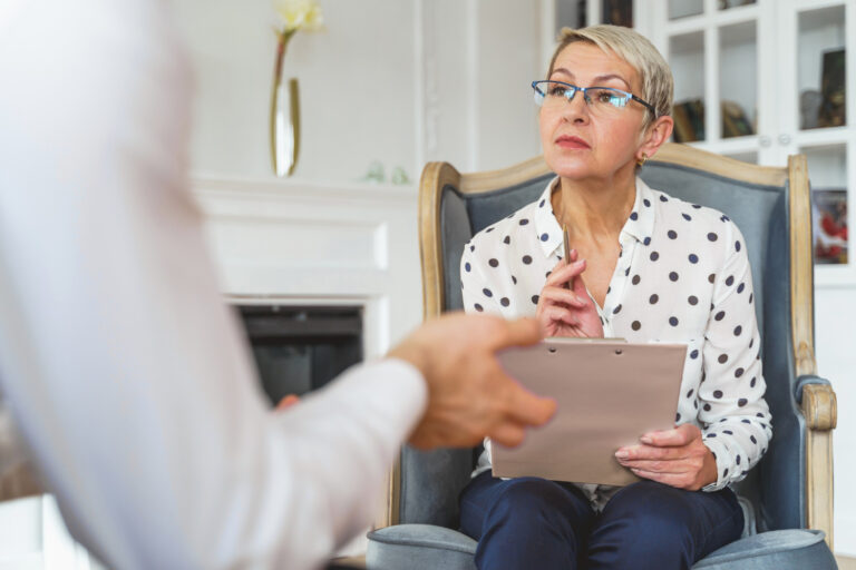 A woman with short blond hair and glasses sits in a chair, holding a clipboard and pen, attentively listening during a forensic psychological evaluation with a person whose hands are visible in the foreground. - KindestMind