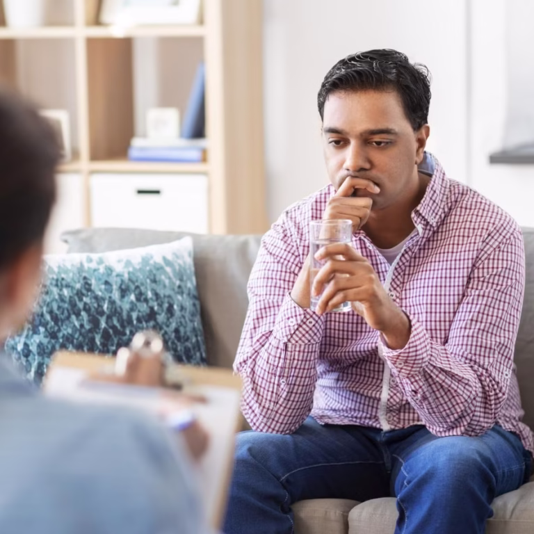 A man sits on a couch, looking thoughtful and concerned, while facing a person holding a clipboard—possibly discussing risk and safety evaluations during a therapy or counseling session in a comfortable, well-lit room. - KindestMind