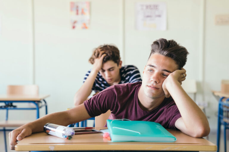 Two teenage students sit at desks in a classroom, looking bored and tired—common ADHD symptoms. One boy rests his head on his hand, while the other has his head down behind him. School supplies are scattered on their desks. - KindestMind