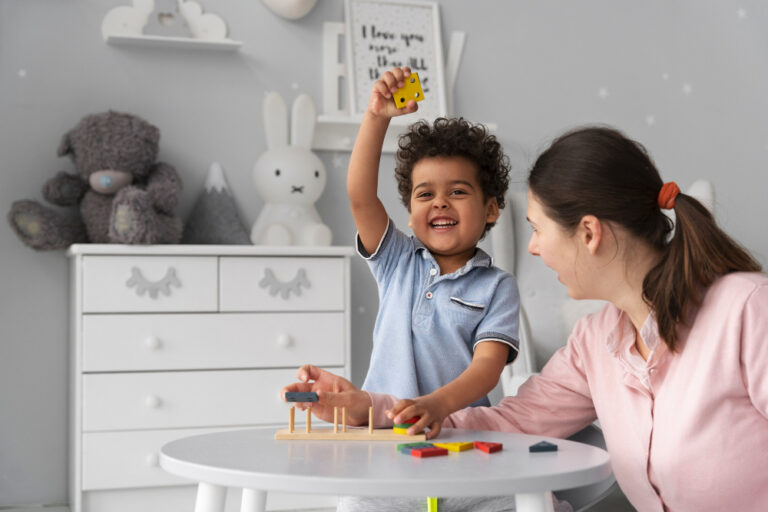 A young child smiles and raises a yellow block excitedly while playing a stacking game with a woman—perhaps during an Autism diagnostic evaluation—in a cozy, gray-themed children’s room decorated with toys and plush animals. - KindestMind