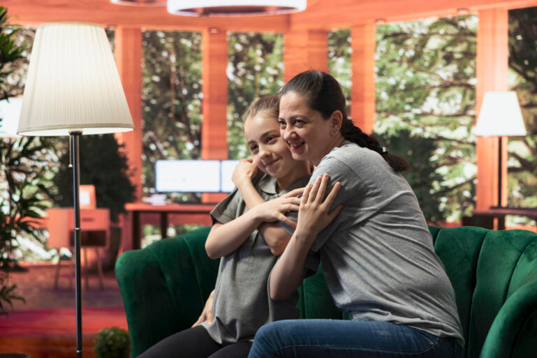 A woman and a young girl, showing autistic traits, sit on a green couch, smiling and hugging each other in a cozy, sunlit room with large windows and lush greenery outside. A floor lamp and a desk with monitors are in the background. - KindestMind