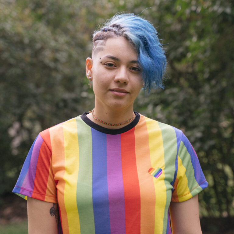 A person with short, blue hair and shaved sides stands outdoors, wearing a rainbow-striped shirt with a small rainbow heart patch—showcasing pride and individuality often embraced in Autism Spectrum Disorder assessments. The background is blurred greenery. - KindestMind