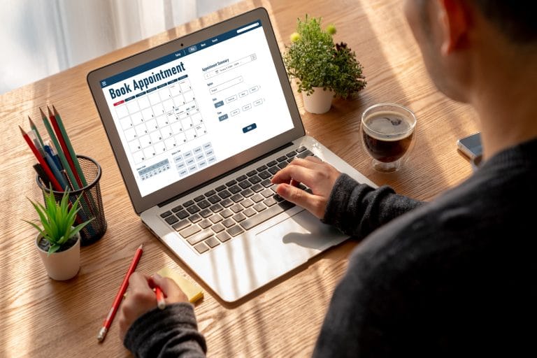 Our Process For Psychological And Neuropsychological Evaluations 3 A person using a laptop to book an appointment online for immigration evaluations, with a calendar on the screen. A cup of coffee, pencil, sticky notes, and small plants are on the wooden desk. - KindestMind