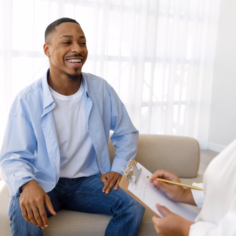 A man sits smiling on a chair, talking with a woman who holds a clipboard and pen, likely conducting a risk assessment, in a bright room with large windows. - KindestMind