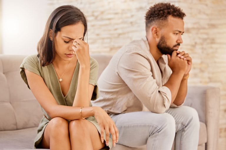 A woman and man sit on a couch looking upset and distant, both appearing stressed and deep in thought. Their body language reflects the emotional challenges they are overcoming together. - KindestMind