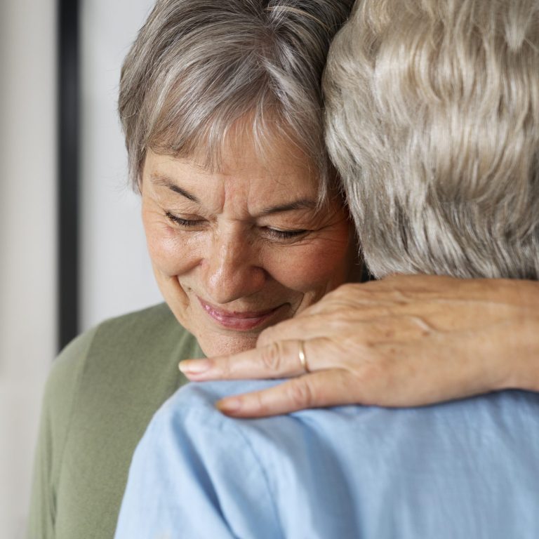 An older woman with short gray hair smiles gently while hugging another person with gray hair, showing the warmth and support often shared by those coping with Dementia or Alzheimer’s. The woman wears a green top; the other person is in a light blue shirt. - KindestMind