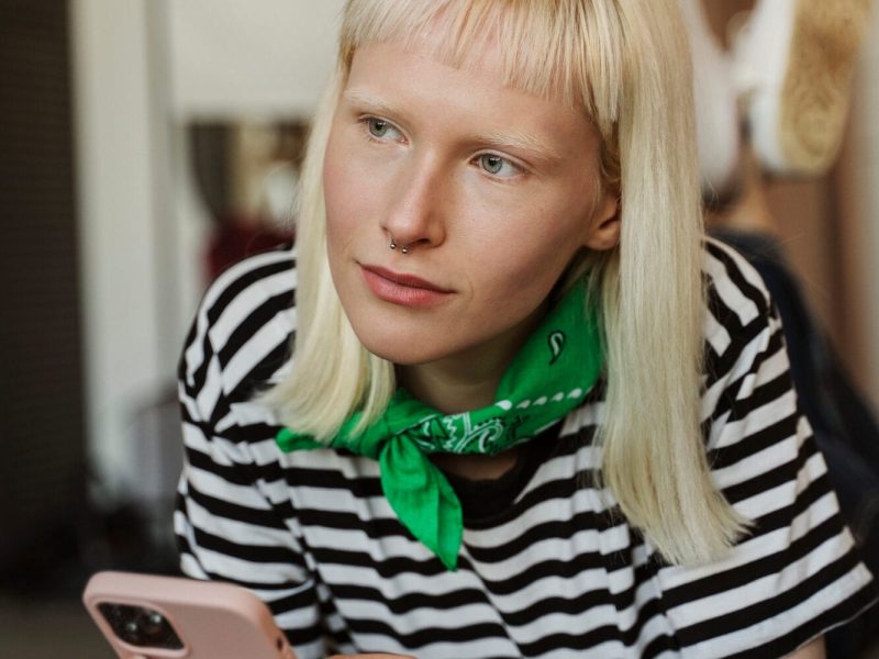 A person with straight blonde hair and a green bandana around their neck holds a smartphone, wearing a black and white striped shirt, and looks thoughtfully into the distance, perhaps reflecting on recent Autism Spectrum Disorder assessments. - KindestMind