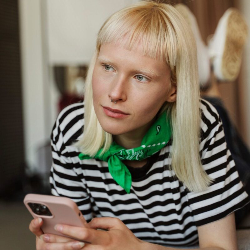 A person with straight blonde hair and a green bandana around their neck holds a smartphone, wearing a black and white striped shirt, and looks thoughtfully into the distance, perhaps reflecting on recent Autism Spectrum Disorder assessments. - KindestMind