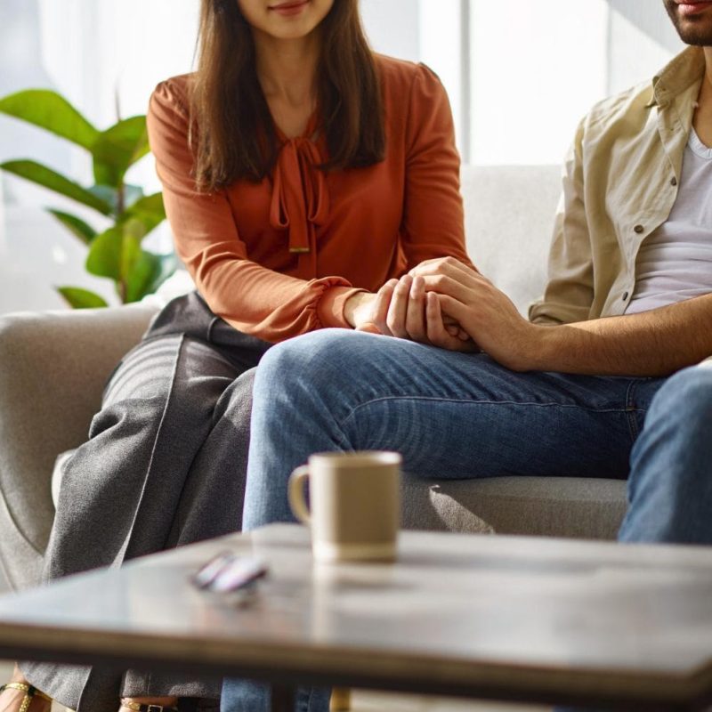 Two people sit close together on a couch. One gently holds the others hand, offering comfort—a quiet moment after discussing Forensic Psychological Evaluations. A coffee mug and glasses rest on a table in the foreground. - KindestMind