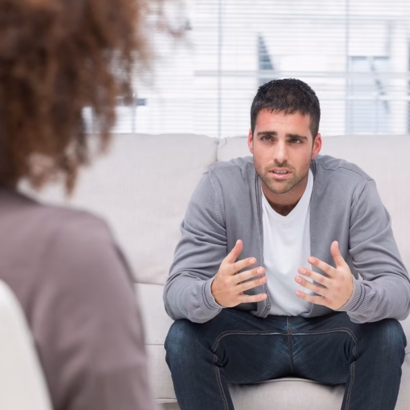 A man sits on a couch, gesturing with concern during what appears to be a counseling session focused on Risk Assessments, while another person listens in the foreground. - KindestMind