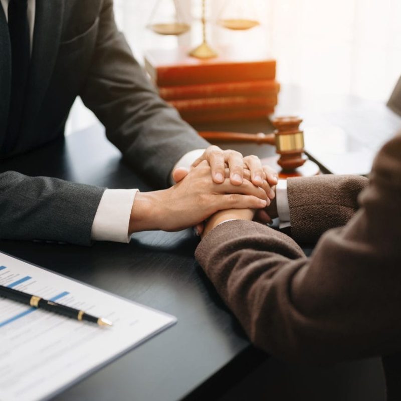 Two people sit at a desk, one offering comfort by placing their hands over the others. A gavel, legal documents, and the KindestMind logo suggest a legal or counseling setting focused on FFDE Psychological Evaluations. - KindestMind