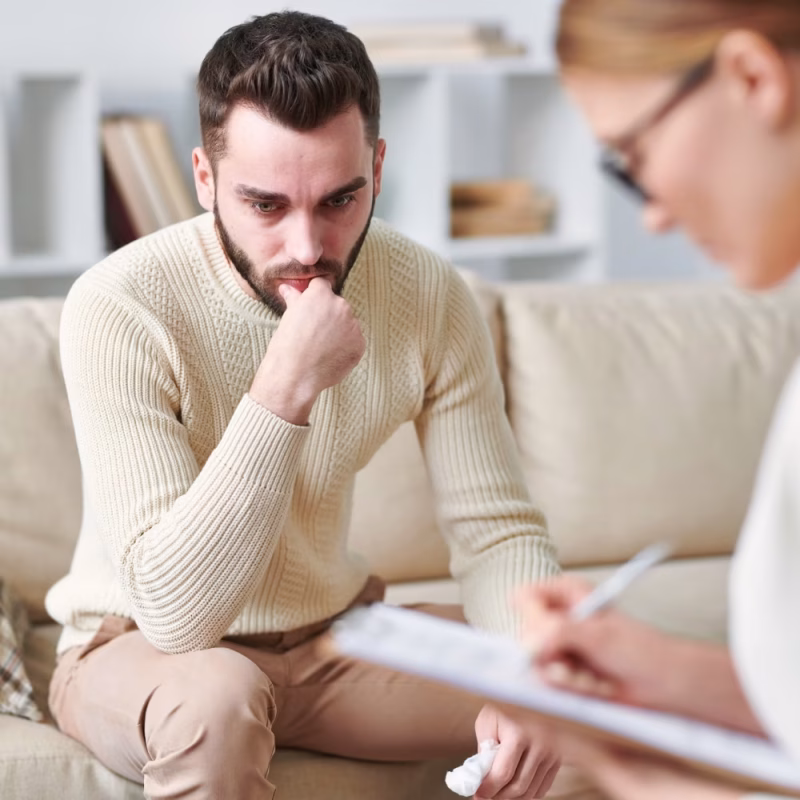 A man sits on a couch, looking pensive with his hand near his mouth, while a woman in glasses conducts a Violence Risk Assessment and writes on a clipboard during a counseling session. - KindestMind