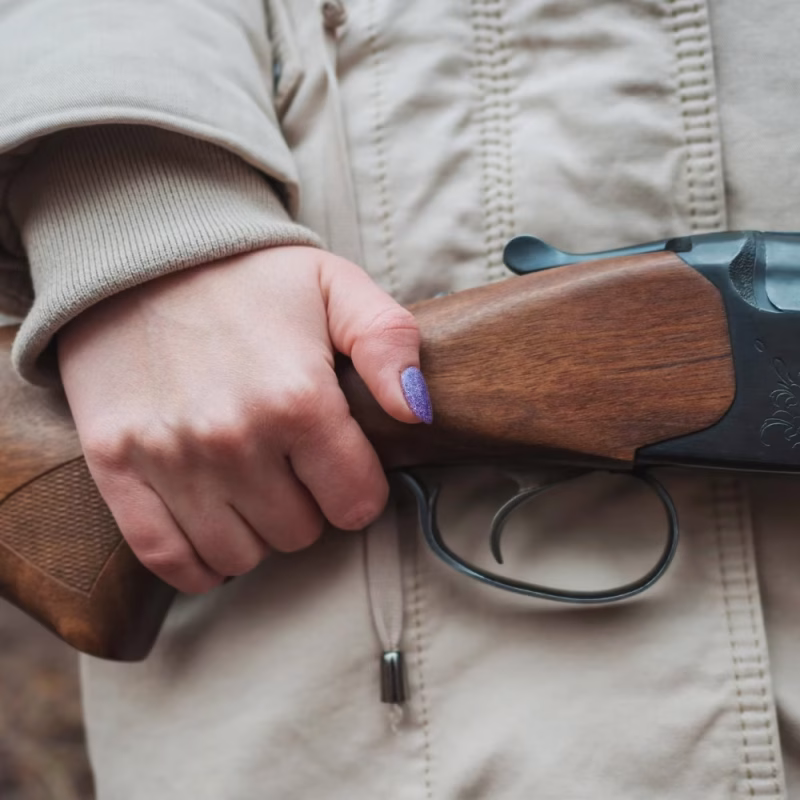 A person wearing a beige jacket holds a shotgun with one hand, showing their purple-painted nails. The background is blurred, suggesting an outdoor setting—perhaps relevant for Capacity to Consent Evaluations in unique environments. - KindestMind