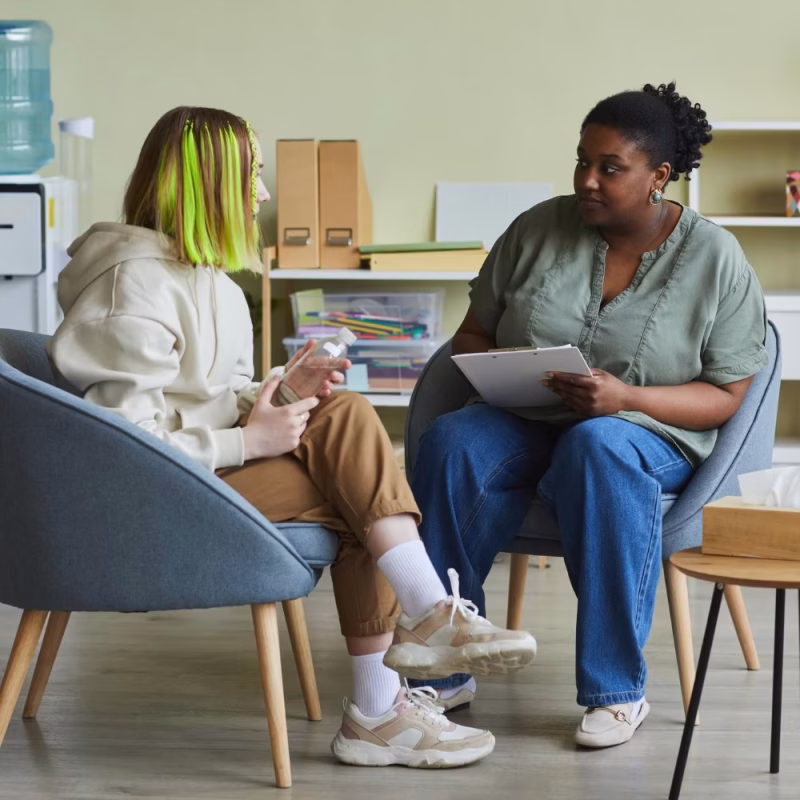 Two people sit facing each other in an office, one holding a clipboard and pen, listening attentively while the other gestures with their hands—an environment typical for Juvenile Justice Evaluations. Shelves and a water cooler are visible in the background. - KindestMind
