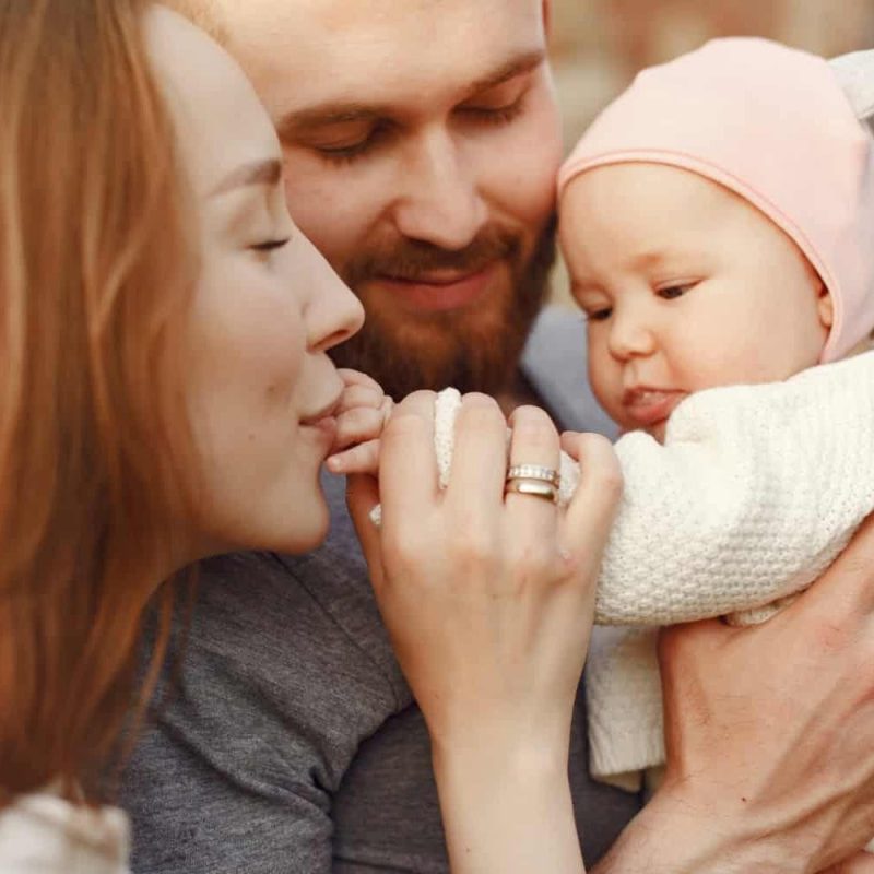 A woman gently kisses a babys hand while a man, smiling, holds the baby close. The baby wears a pink hat and white sweater. The scene captures a tender family moment made possible through Assisted Reproductive Technologies (ART). - KindestMind
