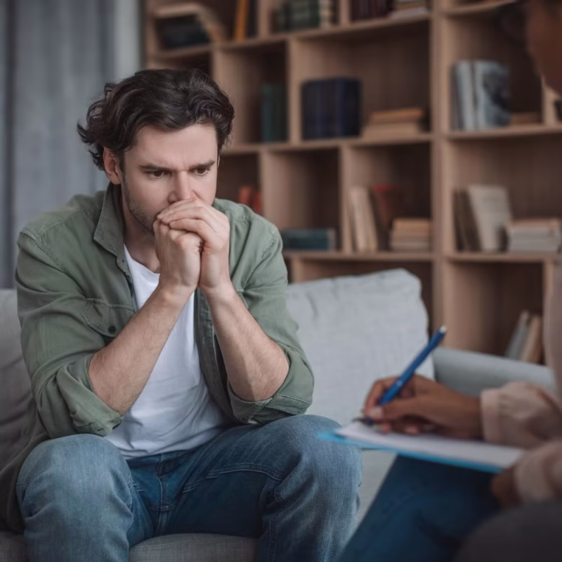 A worried man sits on a couch with his hands clasped near his mouth, looking down, while another person takes notes on a clipboard, suggesting a therapy session focused on risk and safety evaluations in a cozy room with bookshelves. - KindestMind