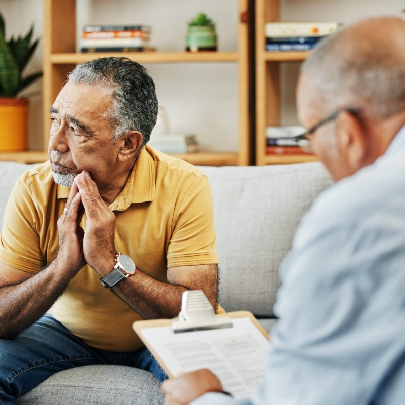 An older man in a yellow shirt sits on a couch looking concerned, his hands clasped near his face, while another person takes notes during psychological evaluations for U-Visa applicants in a cozy, book-filled room. - KindestMind