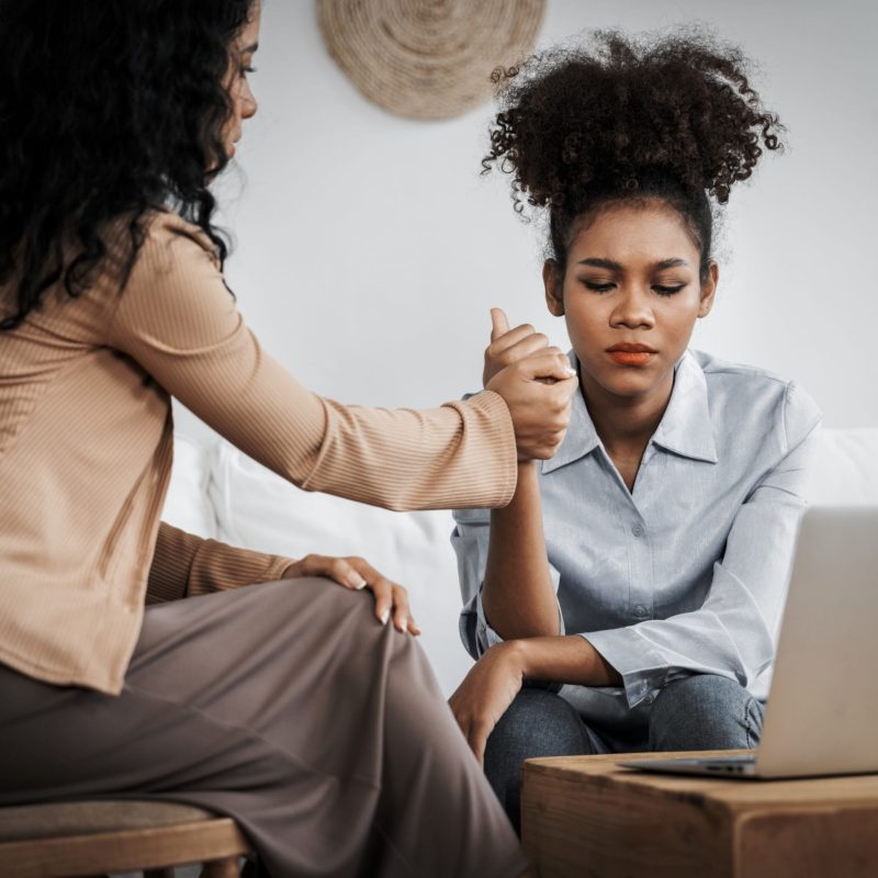 Two women sit together on a couch; one offers comfort by holding the others hand. The woman being comforted, possibly an immigration psychological evaluations or VAWA applicant, looks upset as a laptop sits open on a nearby table. - KindestMind