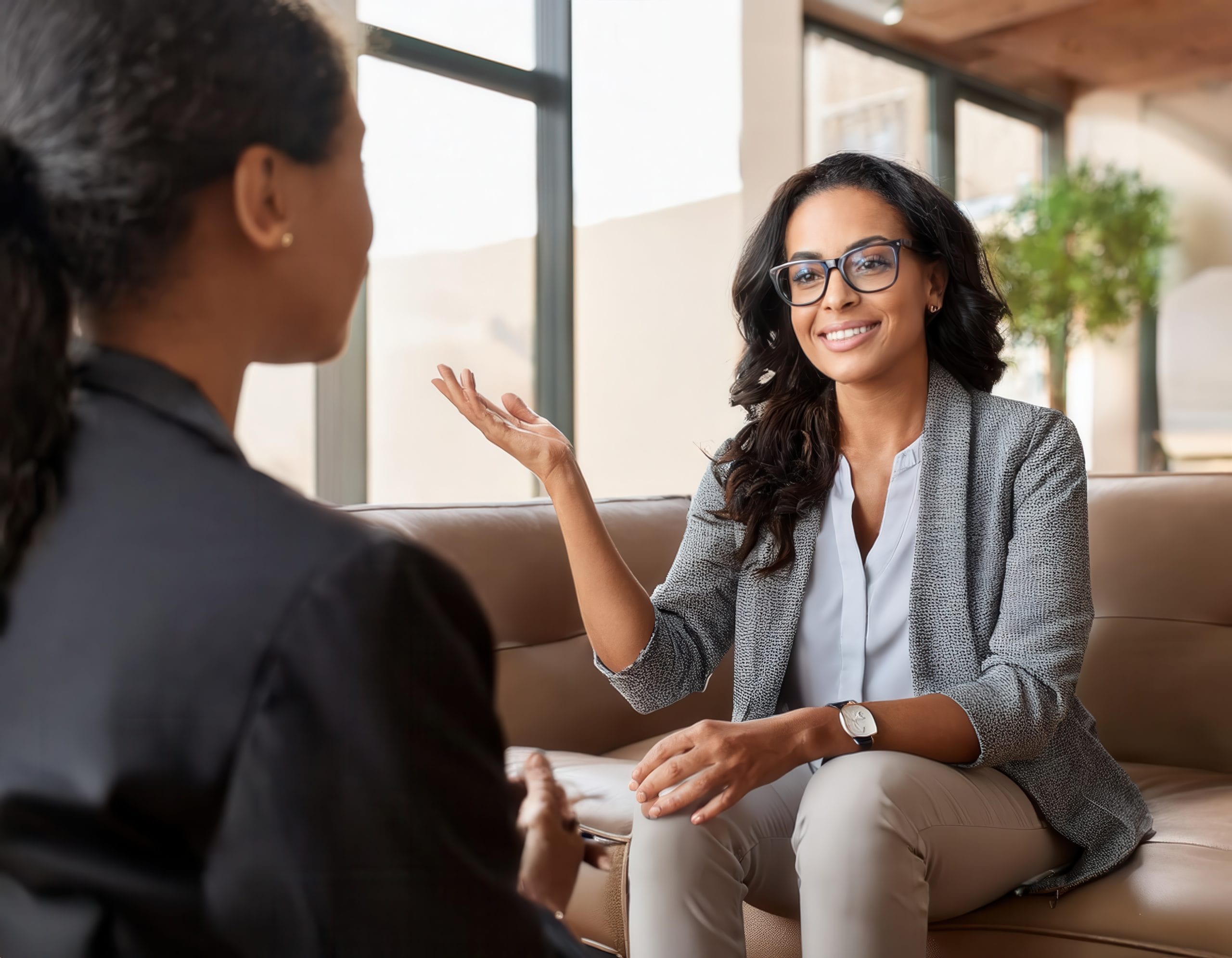 Two women are sitting on a couch in a bright office, engaged in conversation. One woman, smiling and wearing glasses, gestures with her hand while the other listens attentively with her back to the camera. - KindestMind
