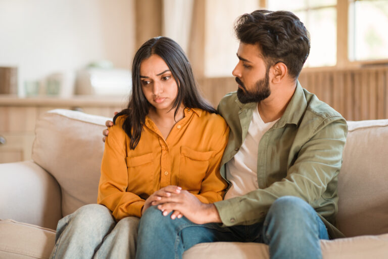 A man sits beside a woman on a couch, gently holding her hand and shoulder. The woman looks down with a sad expression, while the man offers comfort—perhaps awaiting news related to an immigration waiver or extreme hardship waiver. - KindestMind