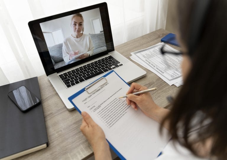 Our Process For Psychological And Neuropsychological Evaluations 4 A person fills out a form on a clipboard while having a video call about the immigration process with a woman on a laptop. Papers, a phone, and a notebook are on the wooden desk in a well-lit room. - KindestMind