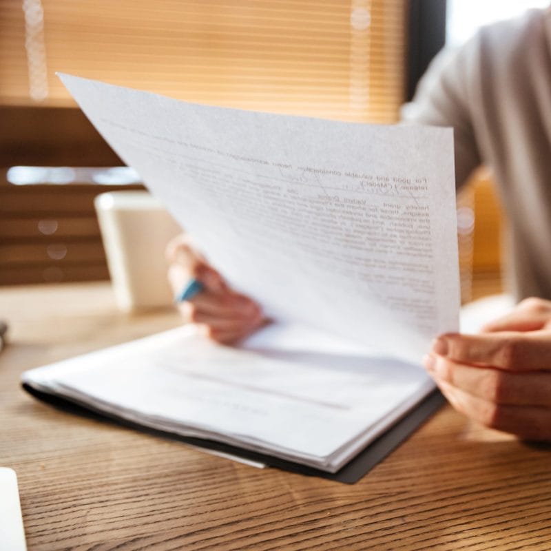 Person sitting at a wooden desk, reading a stack of papers—possibly immigration evaluations. A coffee cup and closed laptop sit in the background, suggesting the careful work of preparing Immigration Psychological Evaluations for KindestMind. - KindestMind