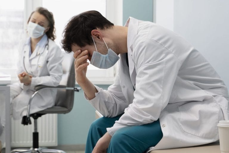 A male medical professional wearing a mask sits with his head in his hand, appearing stressed, while a female doctor in the background looks on with concern—highlighting the pressures faced during Fitness for Duty Evaluations. - KindestMind