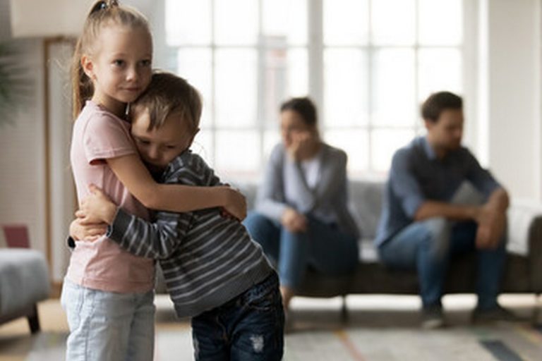 A young girl and boy hug each other in the foreground while two adults, possibly involved in a custody case, sit on a couch in the background looking concerned. Natural light streams through large windows behind them. - KindestMind