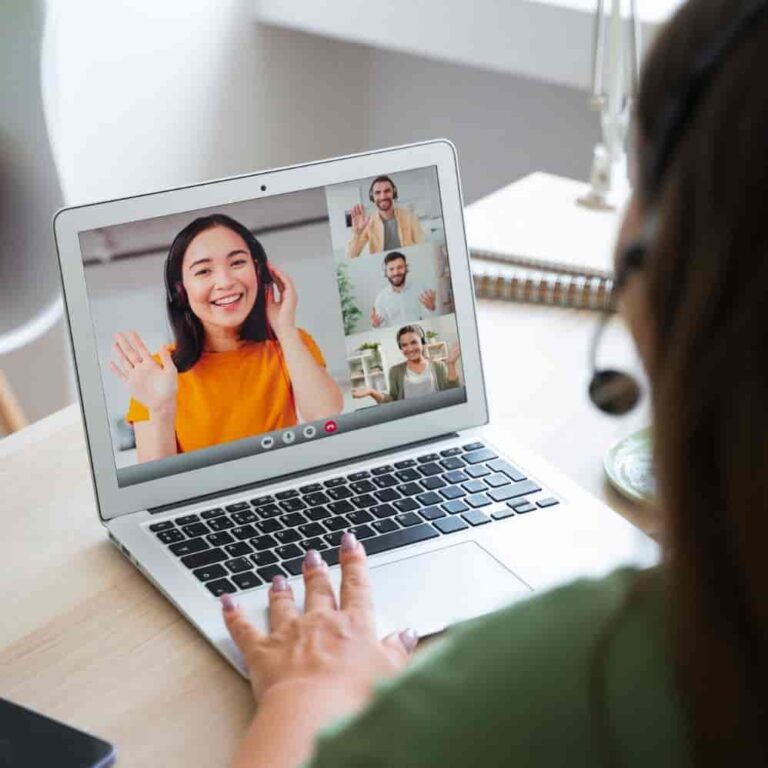 Home 2 Person on a video call using a laptop, waving at three people on the screen, each in their own video feed. The scene suggests a virtual meeting or online chat, with notebooks and a pen nearby on the desk. - KindestMind