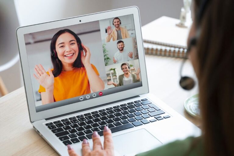 Home 1 A person wearing a headset participates in a video call on a laptop, greeting three smiling people on the screen, each waving from their own locations. A notebook and plants are visible on the desk. - KindestMind