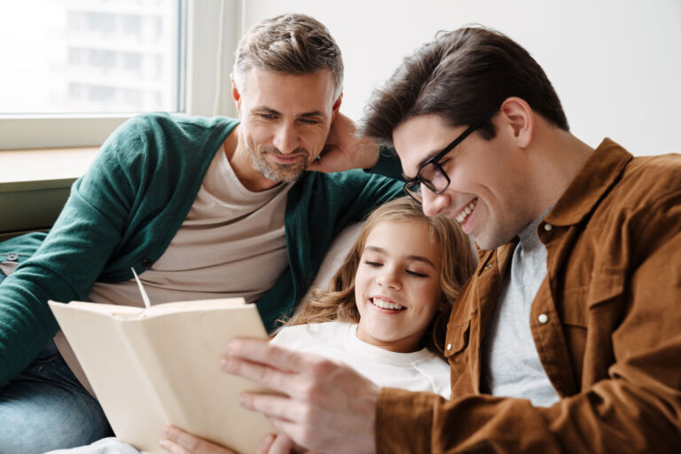 Two men and a young girl, brought together through international adoption, smile and read a book together while sitting on a couch by a window. - KindestMind